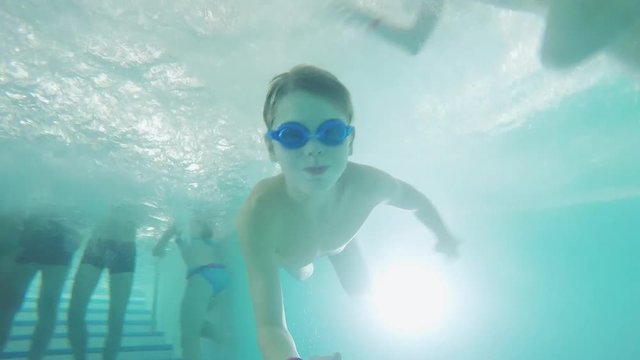 Boy swims under water in pool with swimming goggles through legs other people. 1920x1080