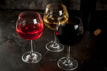 Selection of three kinds (Varieties) of wine - red, white and pink. In glasses, with an open bottle. On a dark concrete background. Copy space