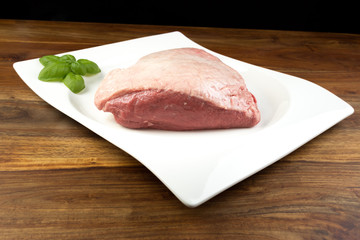 Full piece of raw cow beef (picanha), with basil on a white dish and natural wood table - dark background