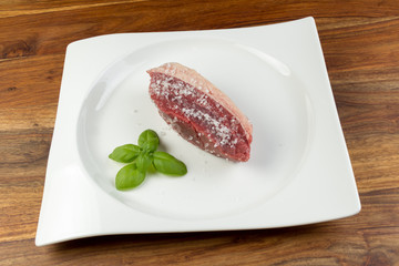 Slice of salted raw cow beef (picanha) and basil leaves on a white dish and vintage natural wood table - coarse salt was used
