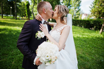 Fashionable kissing wedding couple hugging at green park on sunny wedding day.