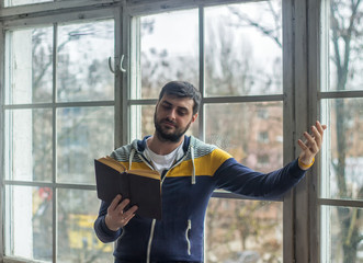 Bearded poet man reads a book with an expression gesture. Big window on background.  - obrazy, fototapety, plakaty
