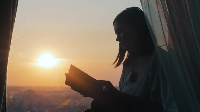 A girl is reading a book on the windowsill at sunset.
