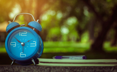 Blue alarm clock and notebook on a wooden desk with blurred nature background.