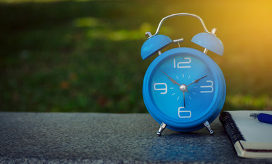 Blue alarm clock and notebook on a wooden desk with blurred nature background.