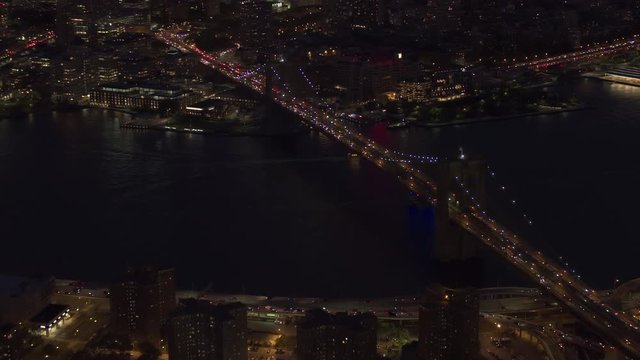 AERIAL HELI SHOT, NO VISIBLE TRADEMARKS: Automobile traffic moving across iconic Brooklyn bridge lit up with lights on magical summer night in New York City. Dense traffic on busy highway and streets