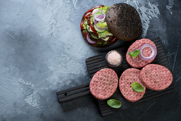 Obraz na płótnie Canvas Raw beef burger cutlets on a black wooden cutting board, high angle view on a grey stone background, copyspace