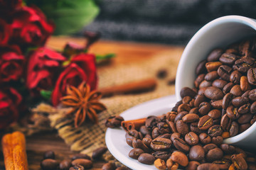 Cup filled with coffee beans on old wooden table with roses in the background.