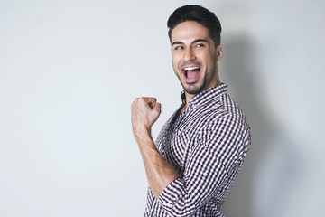 Cheerful young man in checkered shirt smiling and laughing with one fist up, gesturing joy and...