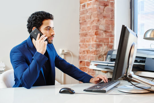 An Arab  businessman in a jacket sitting at a computer and talking on the phone in the office