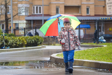 Little boy playing in rainy summer park. Child with colorful rainbow umbrella, waterproof coat and boots jumping in puddle and mud in the rain. Kid walking in autumn shower Outdoor fun by any weather