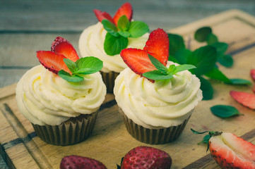 Sweet cupcakes with cream cap,  fresh strawberry and leaves of mint on a wooden background. Vintage toning
