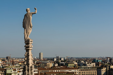 Statues on steeples at the roof of Il Duomo di Milano. Panoramic view on Milano city from the top of the cathedral and antique statue on pillar at background.