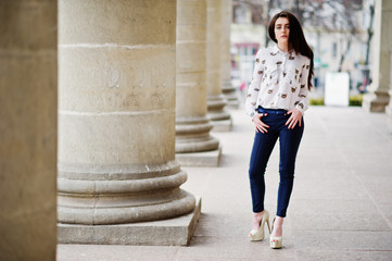 Young stylish brunette girl on shirt, pants and high heels shoes, posed near stone columns. Street fashion model concept.