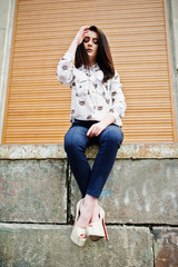 Young stylish brunette girl on shirt, pants and high heels shoes, sitting and posed background orange shutter. Street fashion model concept.