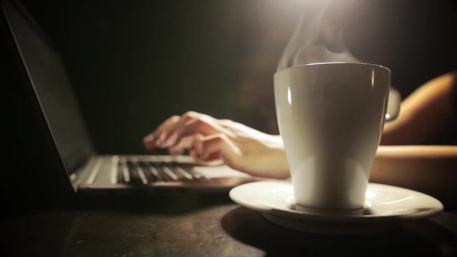 Dolly shot of a woman's hands typing on a laptop keyboard, with a cup of hot coffee near.