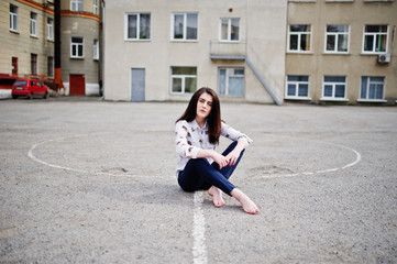 Fototapeta premium Young stylish teenage brunette girl on shirt, pants and high heels shoes, sitting on pavement and posed background school backyard. Street fashion model concept.