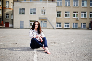 Young stylish teenage brunette girl on shirt, pants and high heels shoes, sitting on pavement and posed background school backyard. Street fashion model concept.