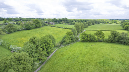 Aerial view of green farm fields, with a hedgerow walking path surrounded by woodlands, in an English countryside, on a cloudy summer day .