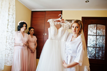 Bride on silk robe looking on her wedding dress against bridesmaids at room on morning day.