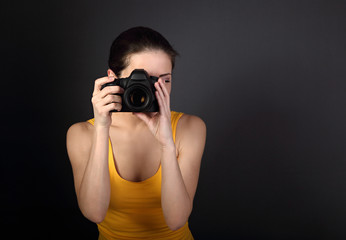 Young female photograph in yellow top holding camera and makeing the photo on dark background with empty copy space. Closeup portrait