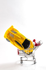 Buying a new car and automobile shopping concept with yellow sports car in a red shopping cart or basket isolated on white