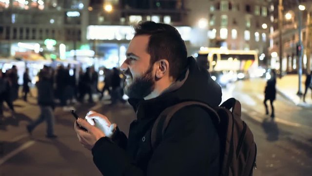 Man Checks His Smartphone Crossing Busy City Intersection At Night