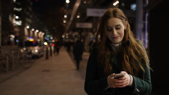 4K Attractive young woman walking through the city at night on her phone, in slow motion