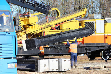 Workers direct a concrete railway pile during unloading by means of a lifting railway crane built into an open freight car