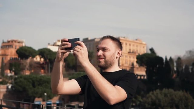 Man tourist explores new city, takes selfie photos of city on smartphone. Male enjoys trip, old buildings. Slow motion.