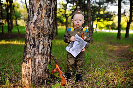 Child boy in military uniform of a Soviet soldier with a photograph of a veteran of World War II in hands against a background of green trees. Victory Day, May 9, against fascism.