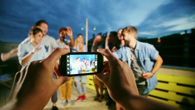 View of hands holding phone and filming happy group of friends dancing on the rooftop terrace in the evening, graded