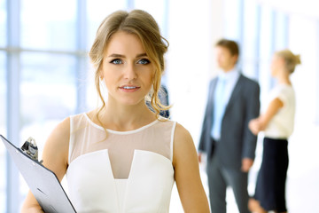 Blonde woman with touchpad computer looking at camera and smiling while business people are shaking hands over office background