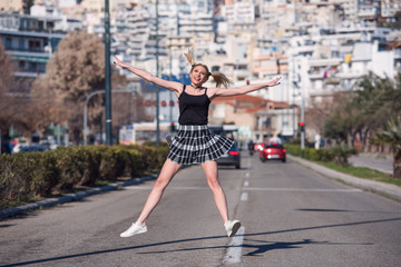 Blond woman with little pigtails and long slim legs wearing mini skirt jump in the middle of a high speed road, cityscape at the background