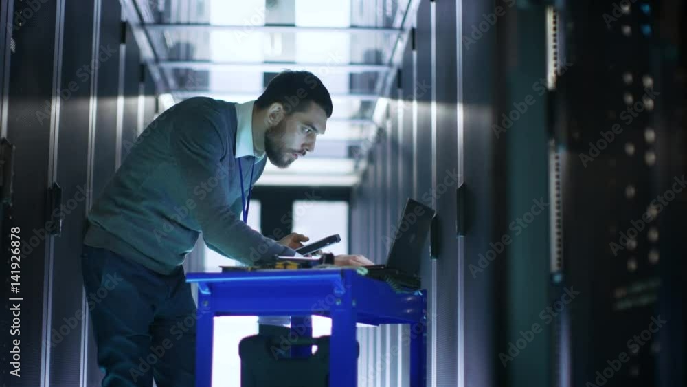 Poster it engineer works on a laptop and scans hard drives that are lying on a crash cart. he works in data