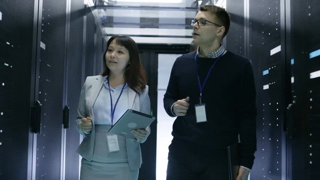 Caucasian Male and Asian Female IT Technicians Walking in Data Center with Rows of Rack Servers. They Have Discussion, She Holds Tablet Computer. Shot on RED EPIC-W 8K Helium Cinema Camera.