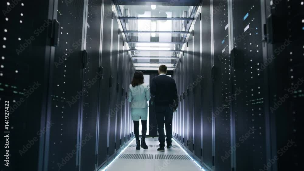 Poster back view of male and female it engineers walking in data center with rows of rack servers. they hav