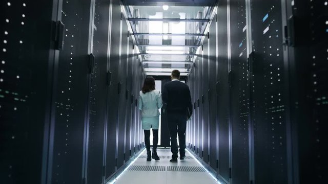 Back View of Male and Female IT Engineers Walking in Data Center with Rows of Rack Servers. They Have Discussion, She Holds Tablet Computer. Shot on RED EPIC-W 8K Helium Cinema Camera.