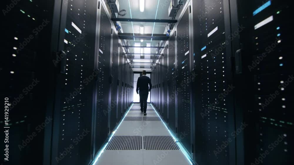 Poster following shot of it engineer walking through data center corridor with rows of rack servers. opens 
