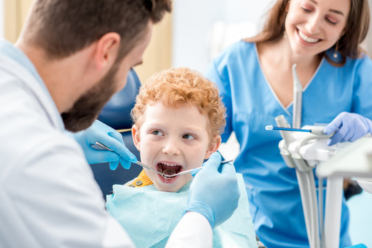 Children's dentist and assistant examinating baby teeth of a young boy sitting on the dental chair at the office