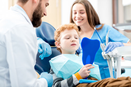 Young boy looking at the mirror with toothy smile sitting on the chair with dentist and assistant at the dental office