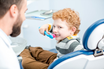 Young boy brushing teeth with big toothbrush sitting on the chair at the dental office