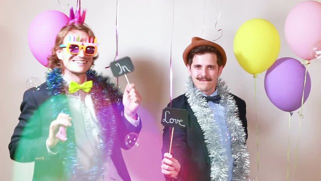 Two funny men holding boards in love in party photo booth, graded in slow motion