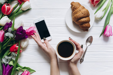 Female hands holding cup of coffee and excellent breakfast of sweet croissant on the smartphone and tulips background for Mother's Day.