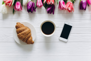 Cup of coffee with croissant, fresh tulips and smartphone on wooden background for Mother's Day.