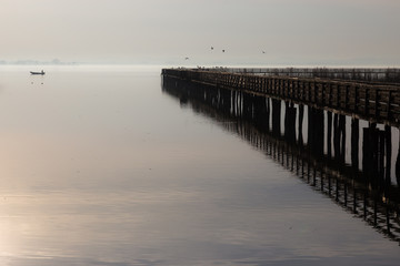 A pier perfectly reflecting on water, with some birds fying, and little fisherman boat at some distance on the opposite side.