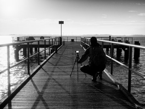 Tall nature photographer at tripod taking picture on the wooden pier in wharf at sunset time.
