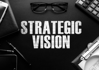 Black chalkboard with business accessories (notepad, magnifying glass, fountain pen, tablet, phone, glasses and calculator) and text STRATEGIC VISION. Top view.
