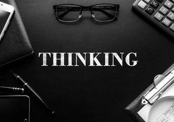 Black chalkboard with business accessories (notepad, magnifying glass, fountain pen, tablet, phone, glasses and calculator) and text THINKING. Top view.