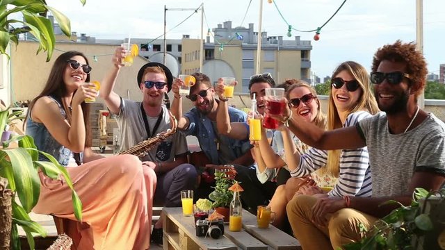 Cheerful multi-ethnic friends looking at camera, laughing and toasting with cocktails at rooftop party on beautiful sunny day
